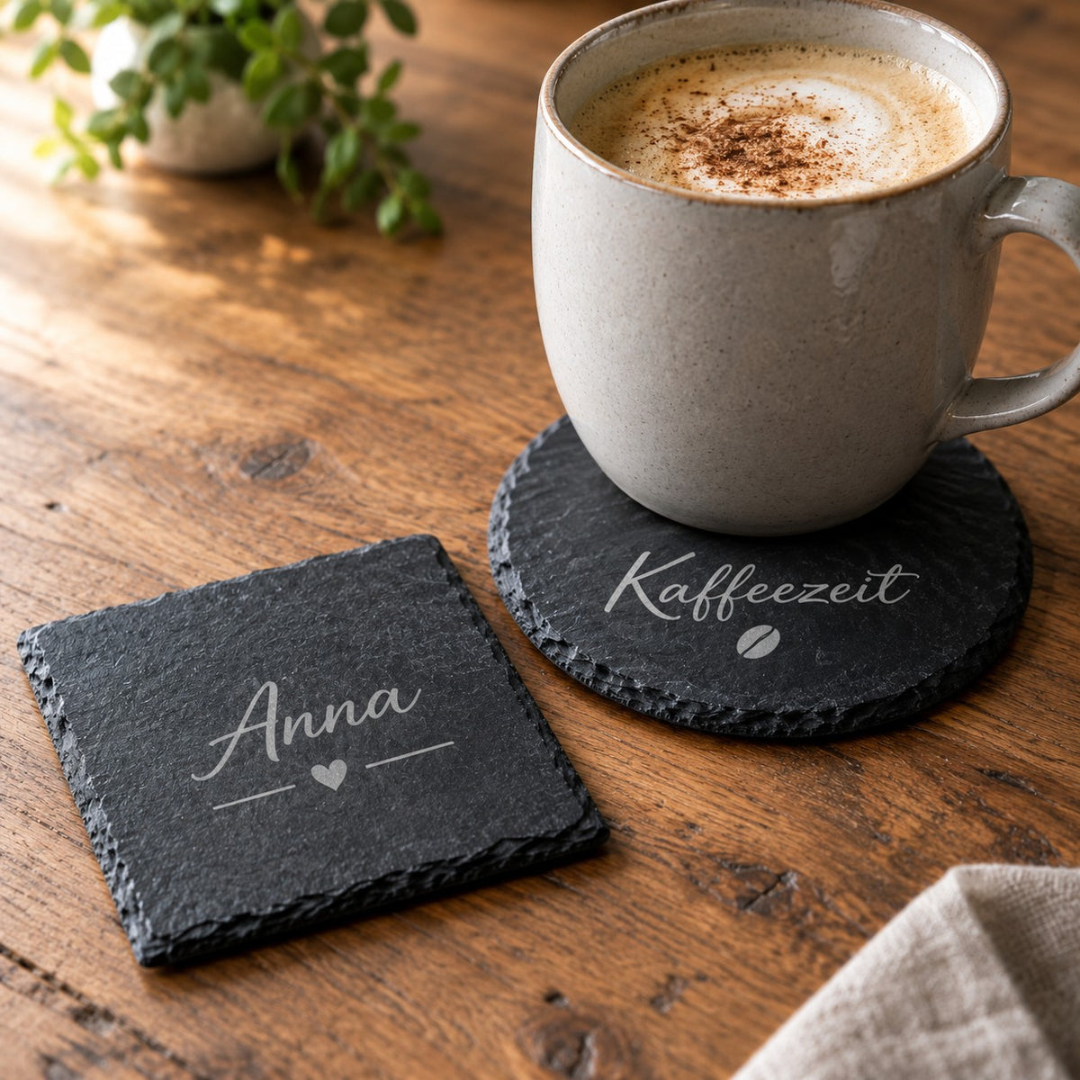 Runder Schiefer-Untersetzer mit Gravur „Kaffeezeit“ unter einer Tasse und eckiger Untersetzer mit Namen „Anna“