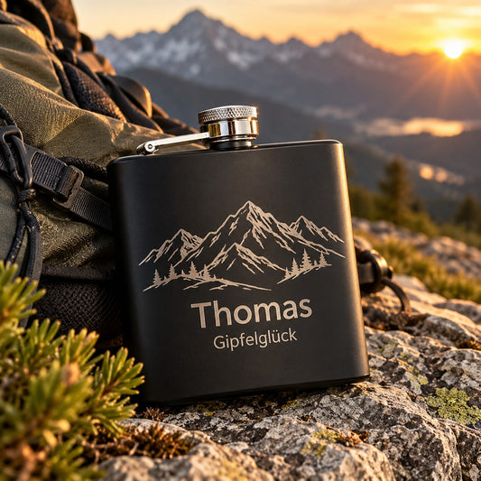 Schwarzer Flachmann mit gravierter Bergkette und Namen „Thomas“ am Wanderrucksack in den Bergen