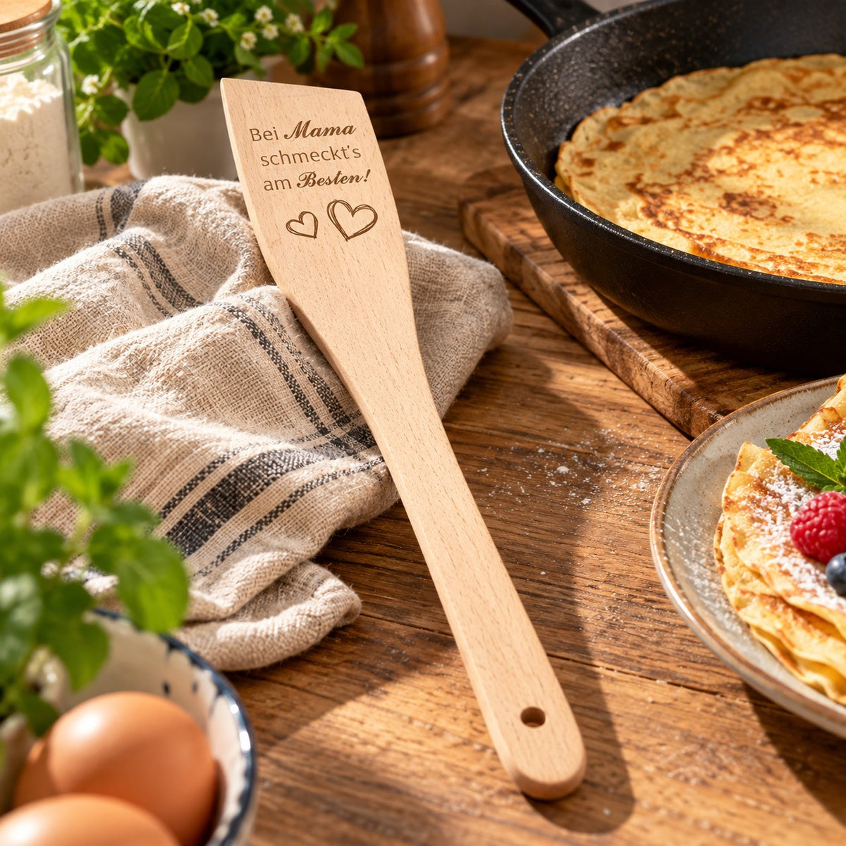 Pfannenwender aus Buchenholz mit Gravur „Bei Mama schmeckt's am Besten!“ und Herzen, neben gusseiserner Pfanne mit Palatschinken auf einer Holz-Arbeitsplatte